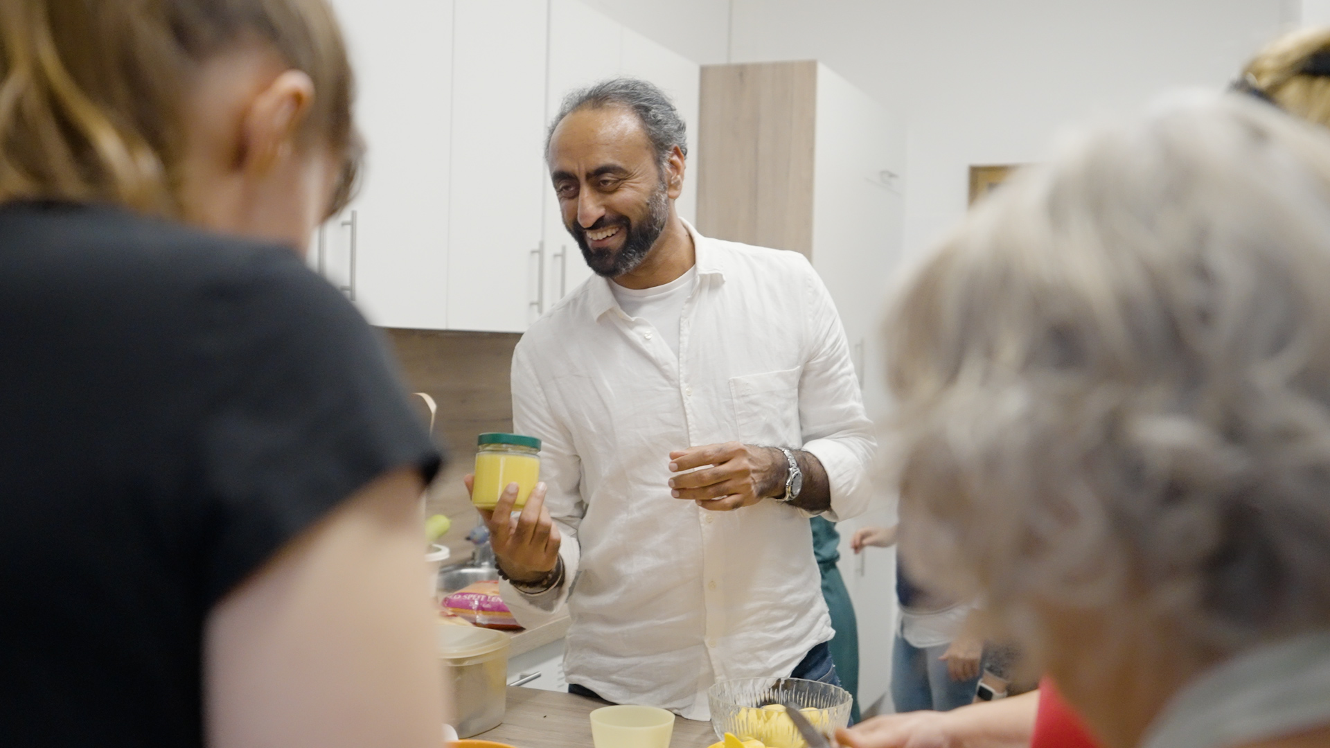 Freiwilligenzentrum Augsburg, kulinarische Weltreise Mann in Küche zeigt anderen Personen ein gefülltes Glas